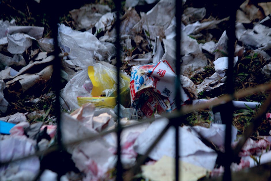 A close-up view of a pile of discarded trash and litter outside, with crumpled paper, plastic bags, and packaging waste scattered on the ground. The debris is seen behind a black metal fence, indicati