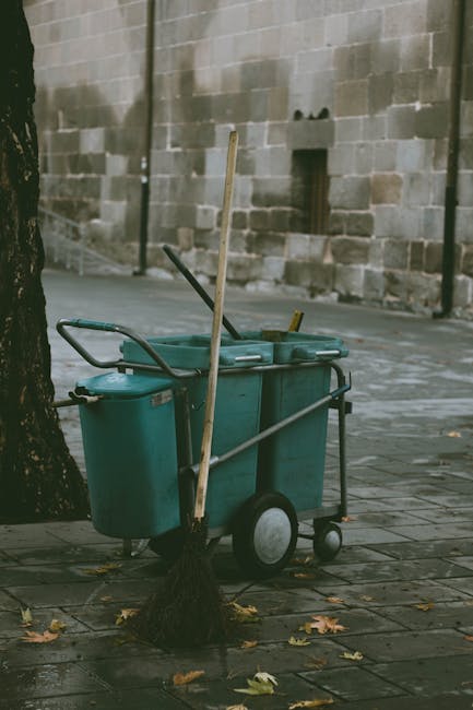 A street scene featuring a large teal-colored waste collection cart with dual wheels, positioned on a paved sidewalk next to a tree. The cart is equipped with a long management pole and a broom with a