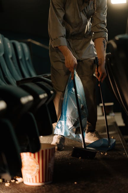 A street scene featuring a large teal-colored waste collection cart with dual wheels, positioned on a paved sidewalk next to a tree. The cart is equipped with a long management pole and a broom with a